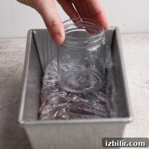 A hand places an empty glass jar inside a plastic wrap-lined metal loaf pan on a light-colored surface.