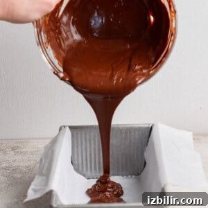 A hand pours melted chocolate from a glass bowl into a parchment-lined rectangular metal baking pan.
