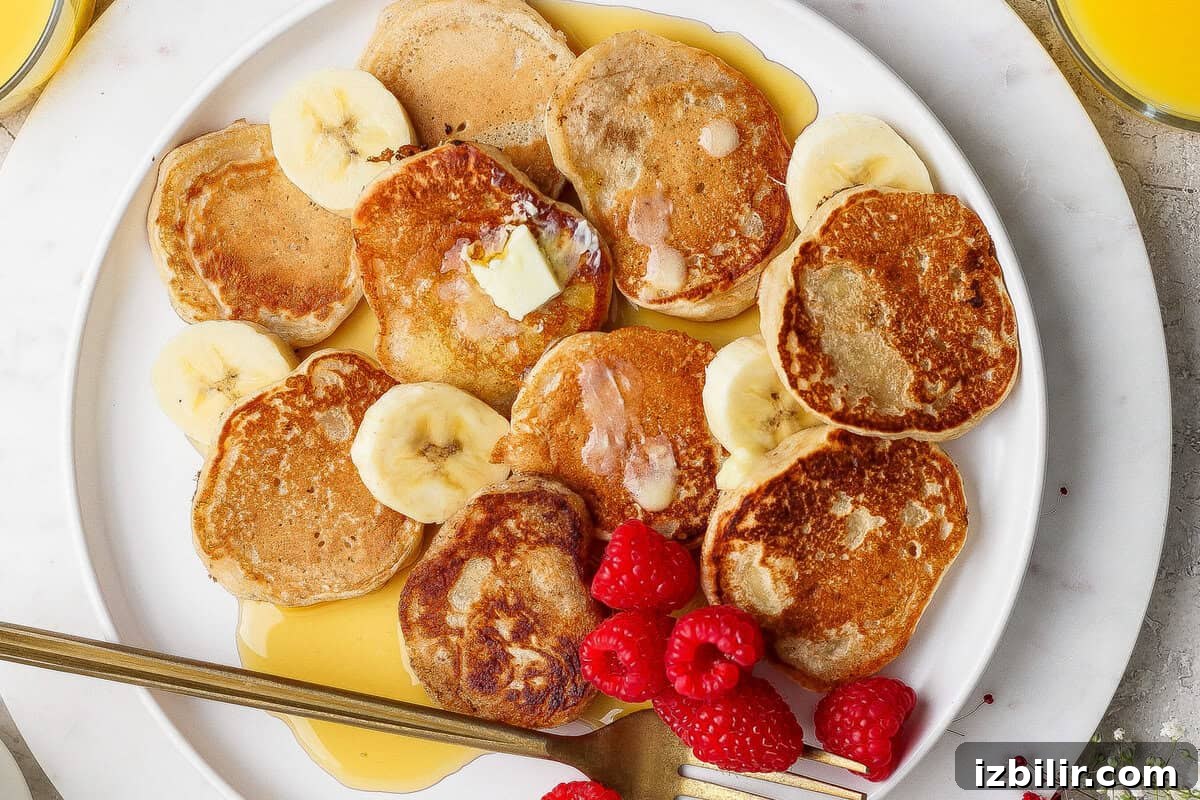 A plate of mini pancakes topped with butter, banana slices, raspberries, and syrup, with a fork on the side.