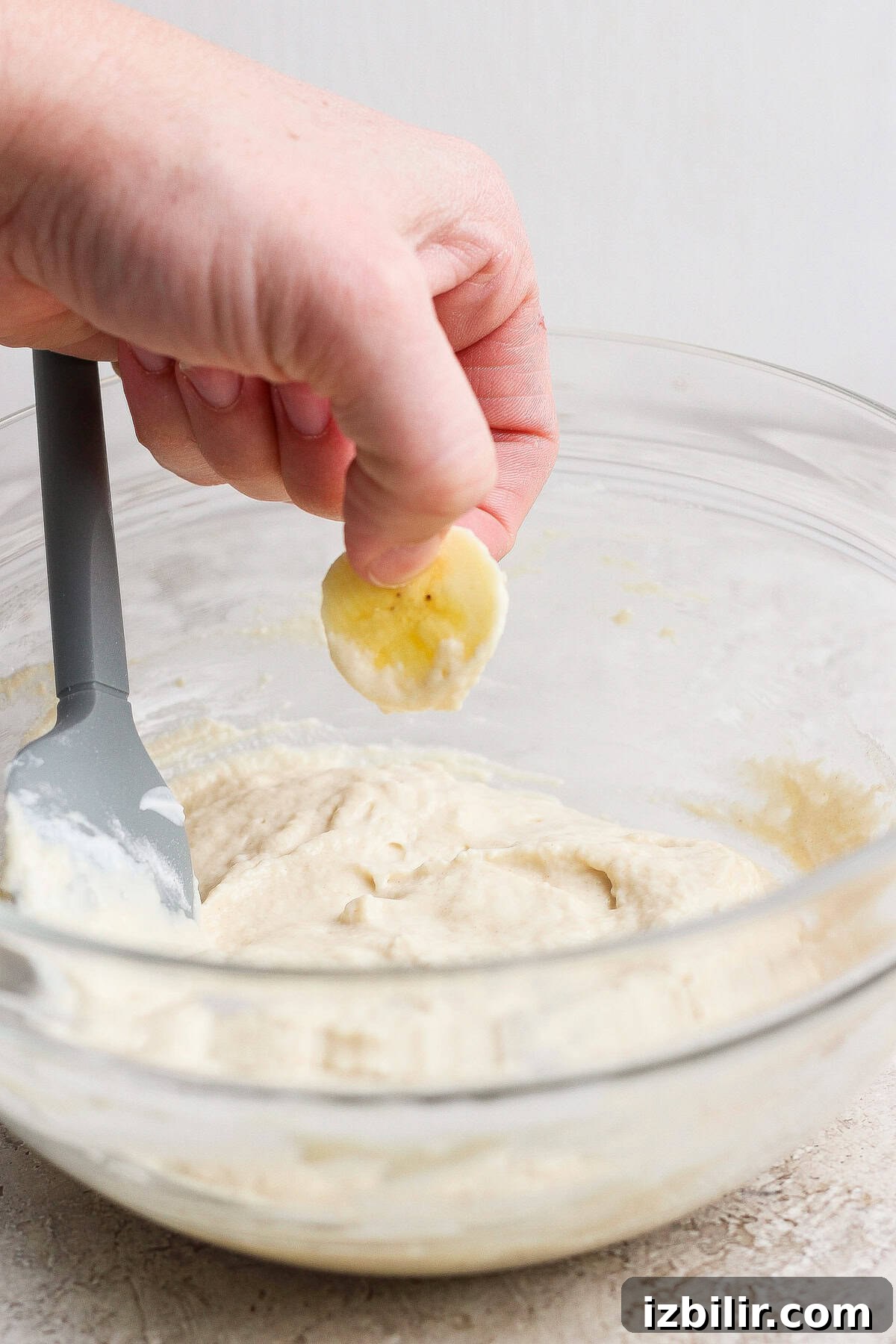 A hand holds a banana slice above a bowl of batter with a spatula resting inside the bowl.
