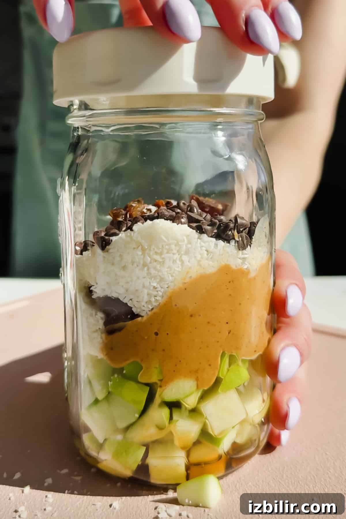 Caramel DeLites Apple Salad in a Jar 3 A hand holds a glass jar filled with chopped green apple, peanut butter, shredded coconut, chocolate chips, and cacao nibs, viewed from the side, with a lid being placed on top.