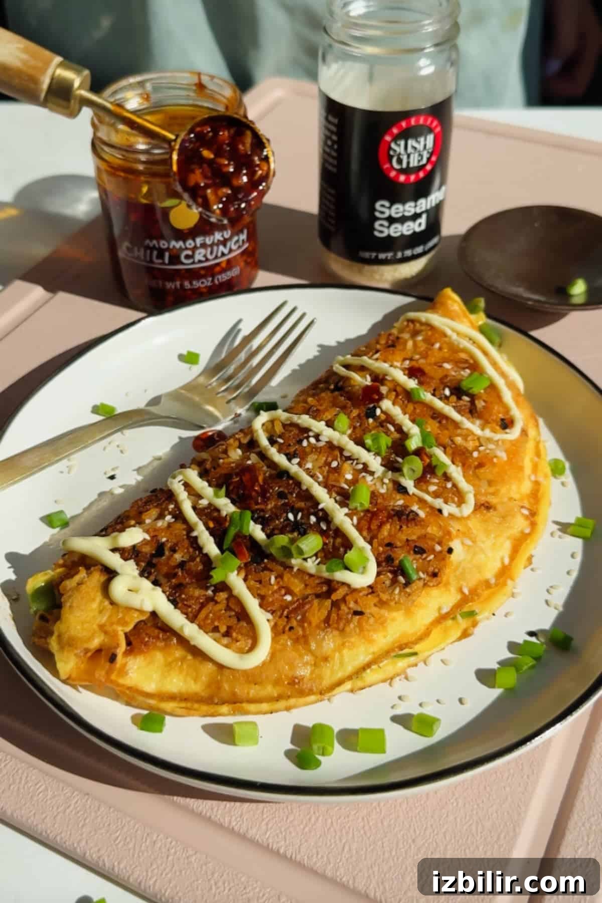 Crispy Fried Rice Omelette Recipe 2 An omelette topped with mayonnaise, chili crunch, sesame seeds, and chopped green onions is served on a white plate with a fork. An open jar of chili crunch and sesame seeds are nearby.