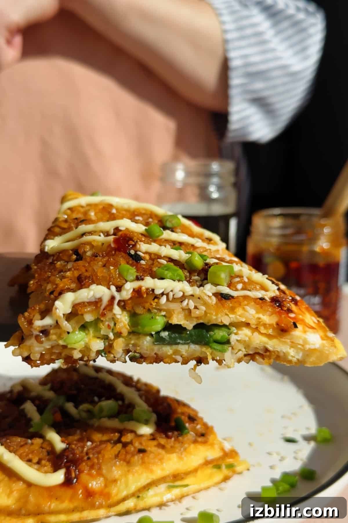 Crispy Fried Rice Omelette Recipe 4 A close-up of a savory slice of food topped with chopped green onions and a drizzle of mayonnaise, being held above a plate with more slices.