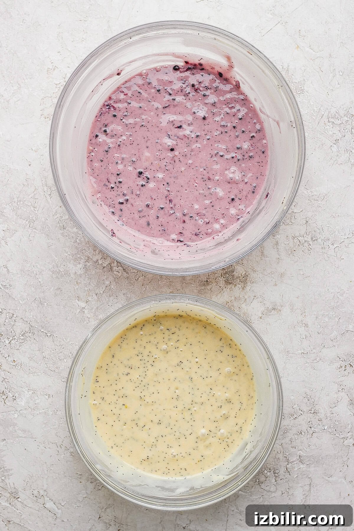 Glazed Blackberry Lemon Poppy Seed Loaf Recipe 3 Two glass bowls on a light surface, one with purple berry-speckled batter and one with yellow batter containing small black seeds.