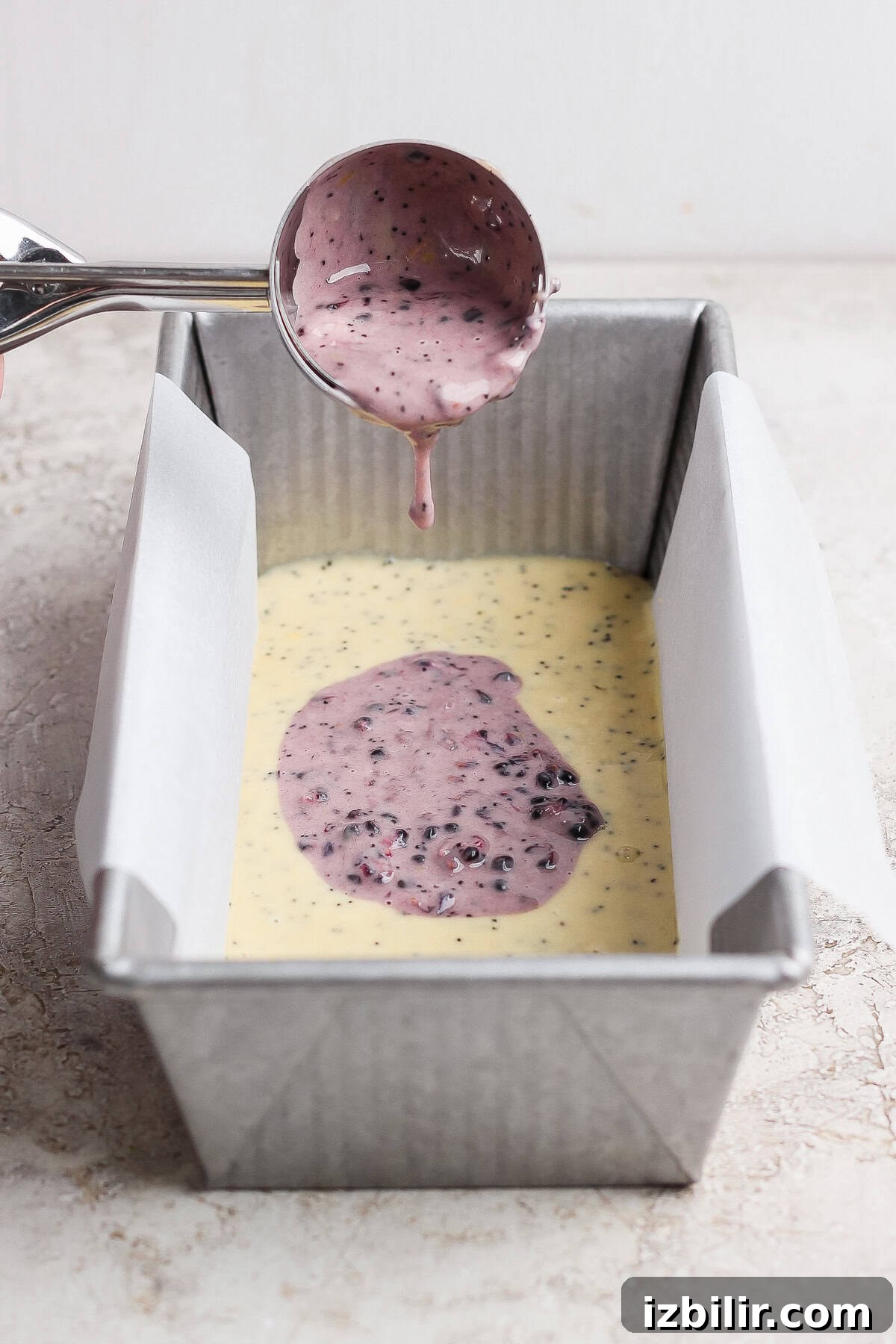 Glazed Blackberry Lemon Poppy Seed Loaf Recipe 4 A metal scoop pours purple berry batter over yellow batter in a parchment-lined loaf pan on a light surface.