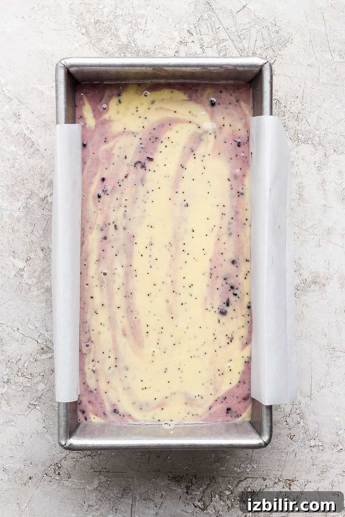 Glazed Blackberry Lemon Poppy Seed Loaf Recipe 5 Rectangular loaf pan lined with parchment paper, filled with swirled yellow and purple cake batter containing small dark specks, on a light textured surface.