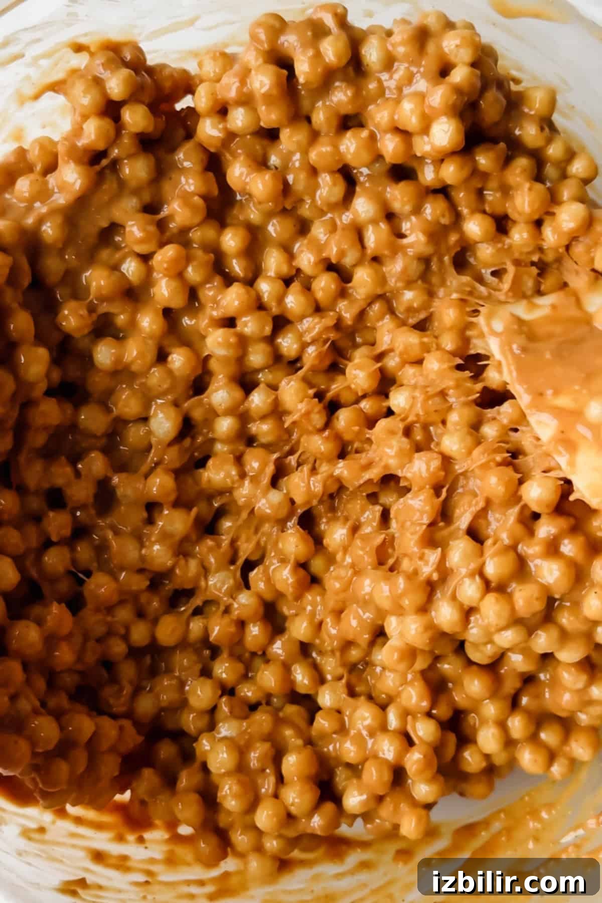 Close-up of a mixture of small round cereal pieces coated in a thick, light brown sauce with a wooden spoon partially visible on the right side.