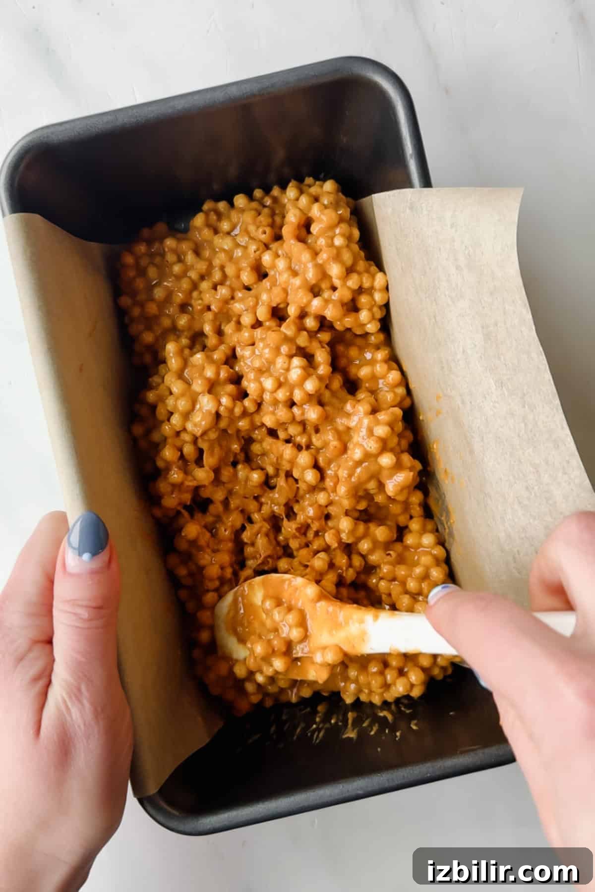 Hands spreading a mixture of coated puffed cereal into a parchment-lined loaf pan with a white spatula.