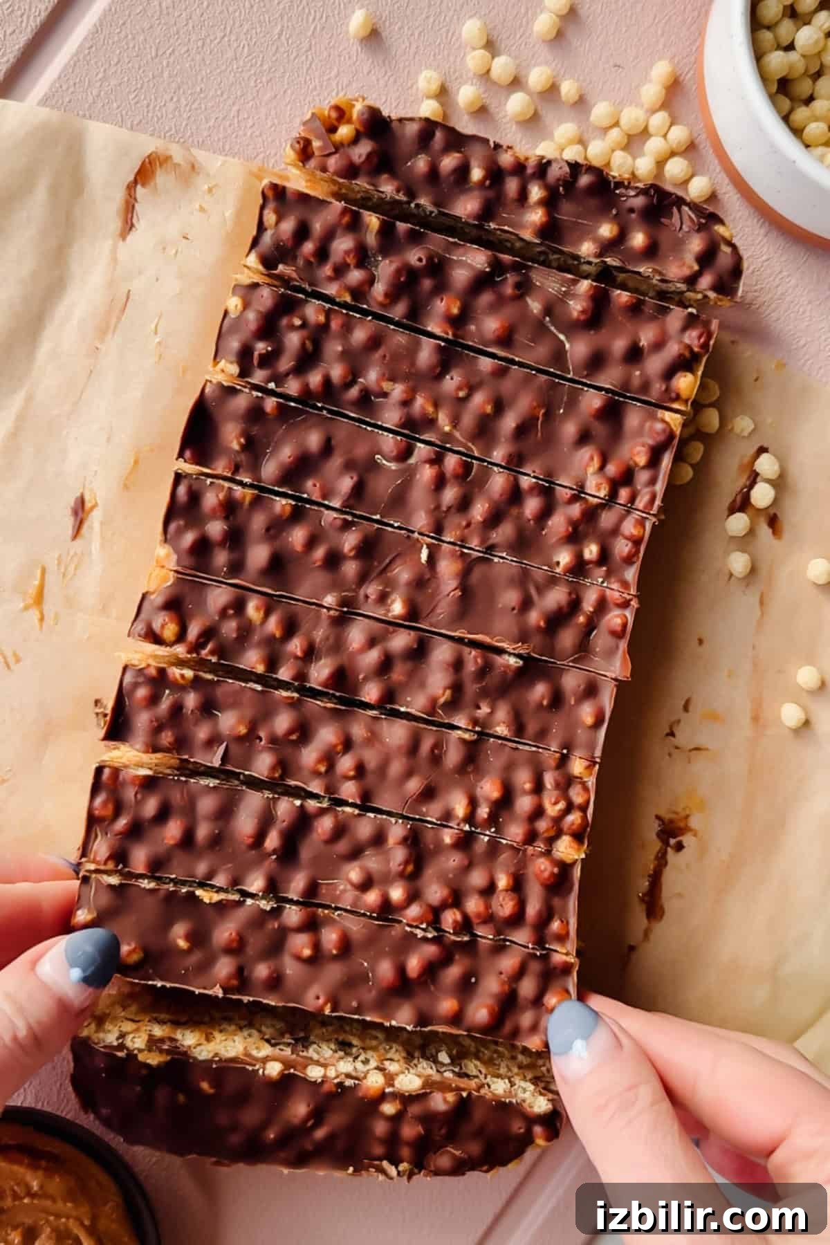 Rectangular chocolate-covered crispy bars with a textured surface, sliced into strips on parchment paper. A person holds one end; puffed grains and a bowl are nearby.