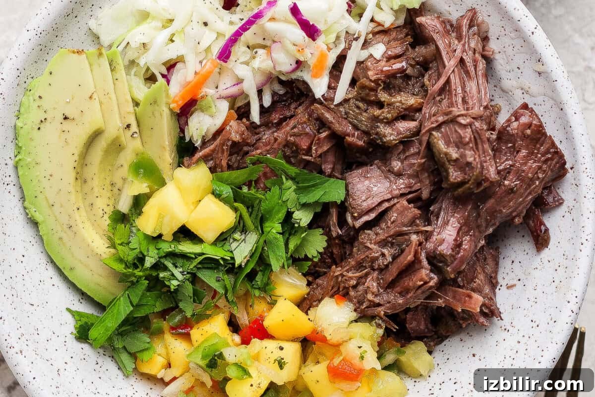 A bowl containing shredded beef, sliced avocado, coleslaw, chopped cilantro, and pineapple salsa.