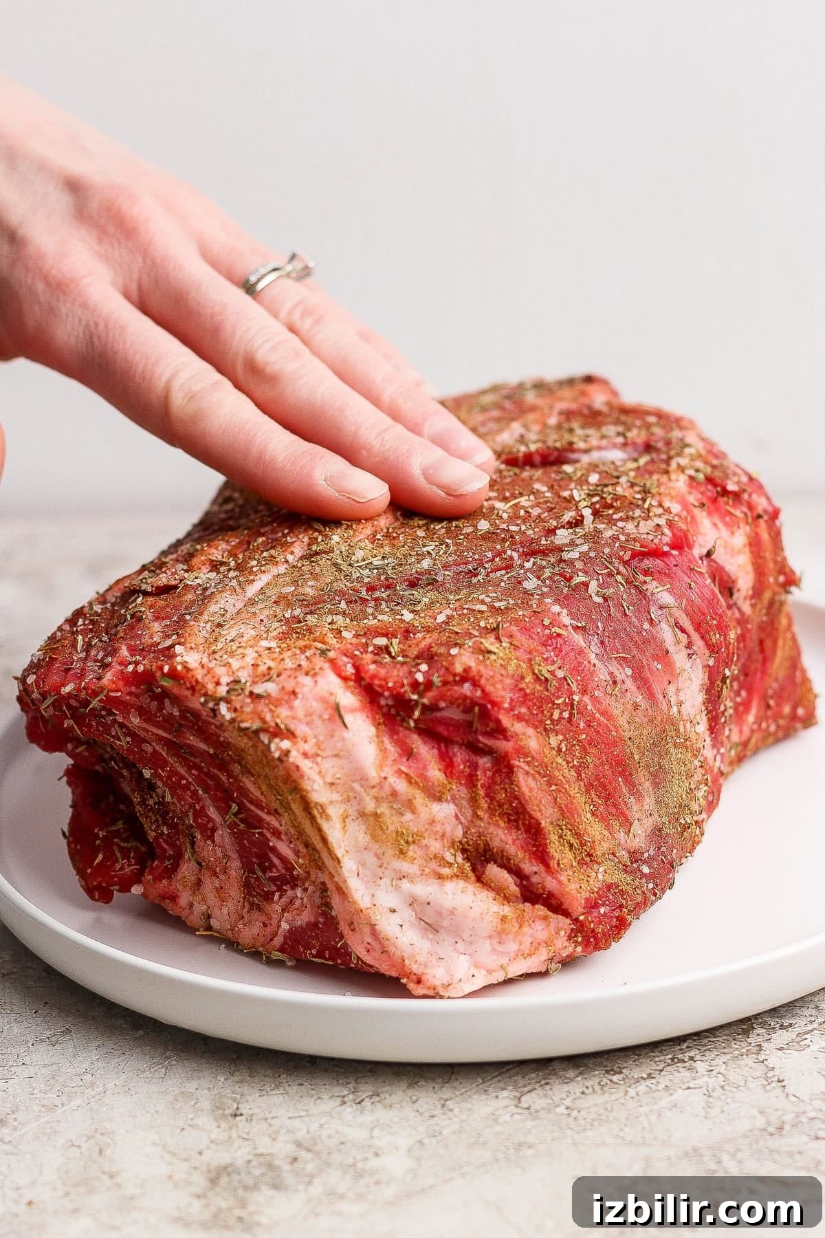 A hand seasoning a raw beef roast with spices and herbs on a white plate.
