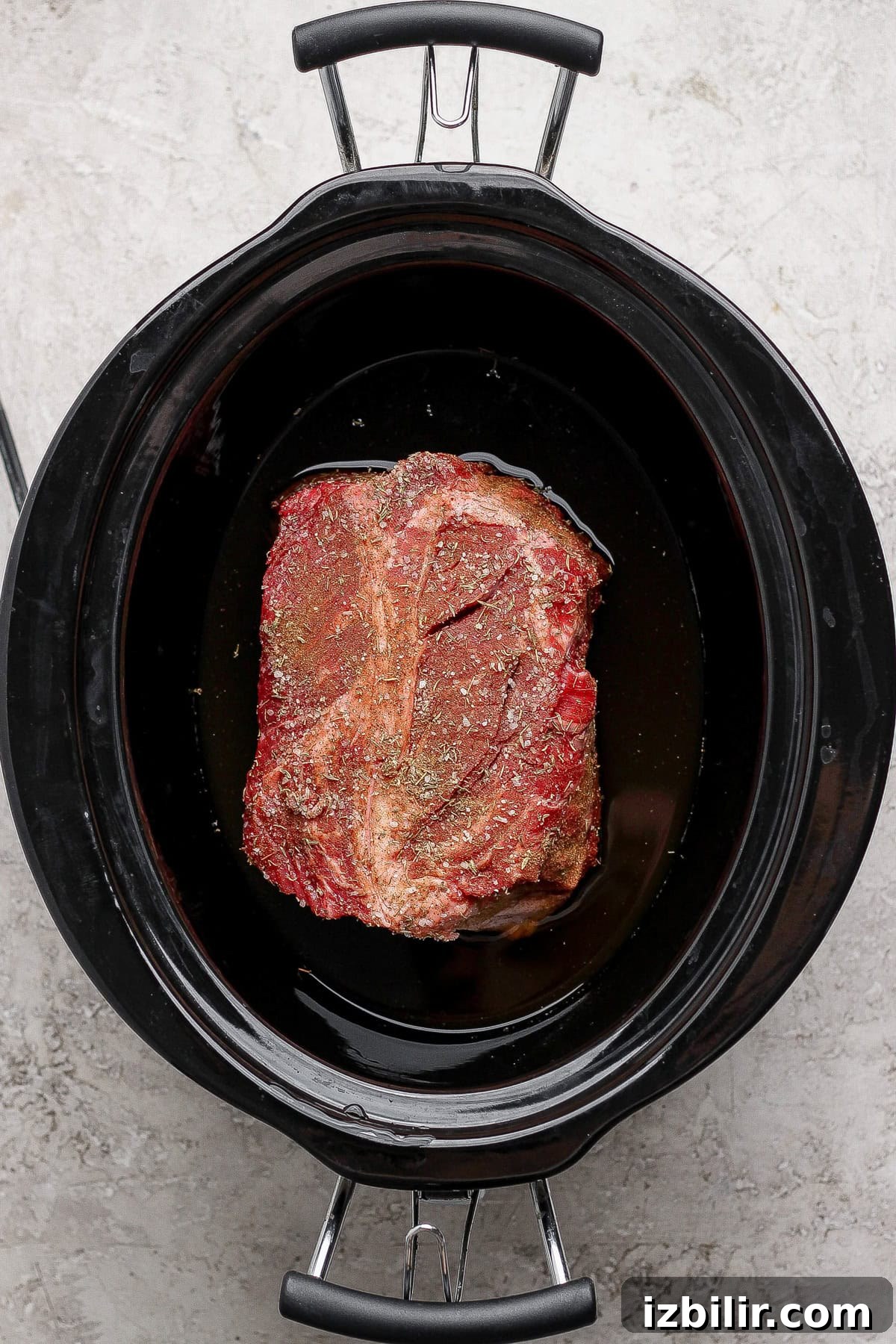 Raw seasoned beef roast sits in a black slow cooker with some liquid at the bottom, viewed from above on a light-colored surface.