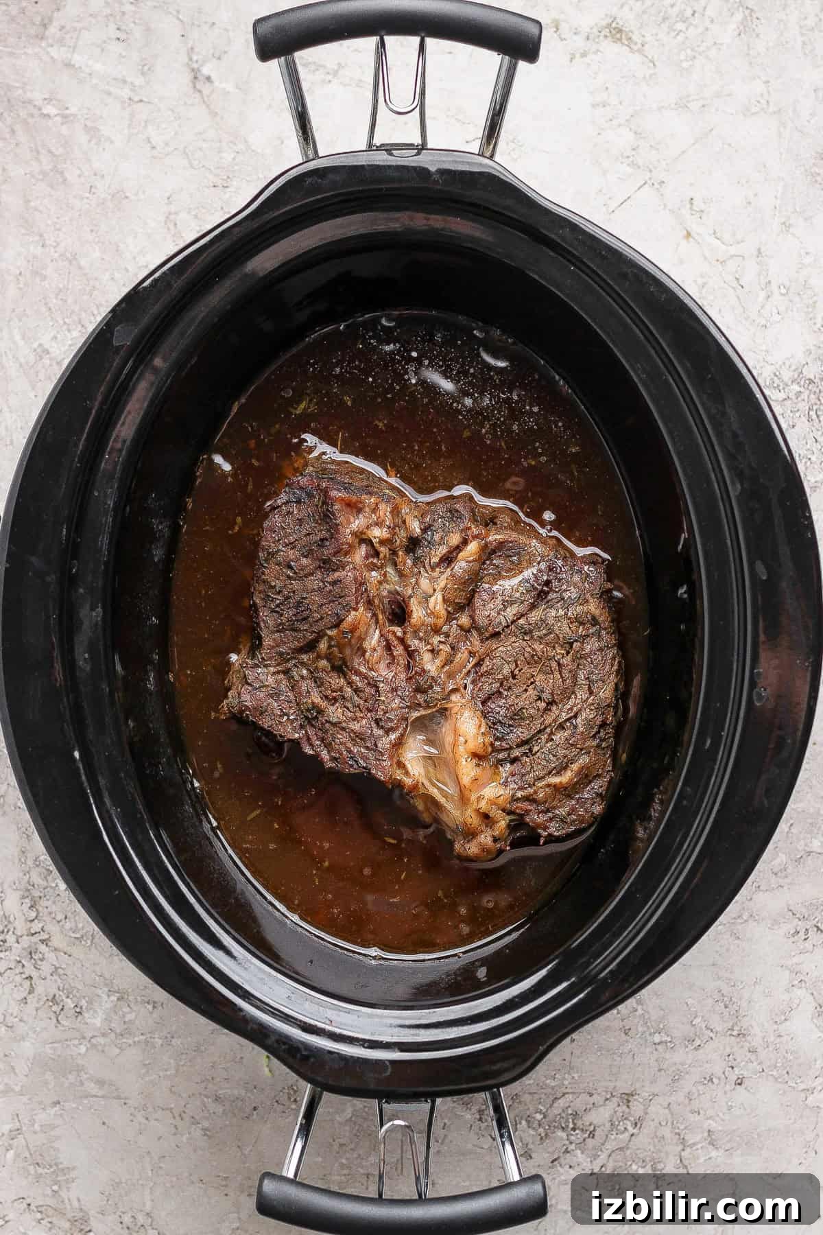 A cooked beef roast sits in broth inside a black slow cooker on a light-colored countertop.