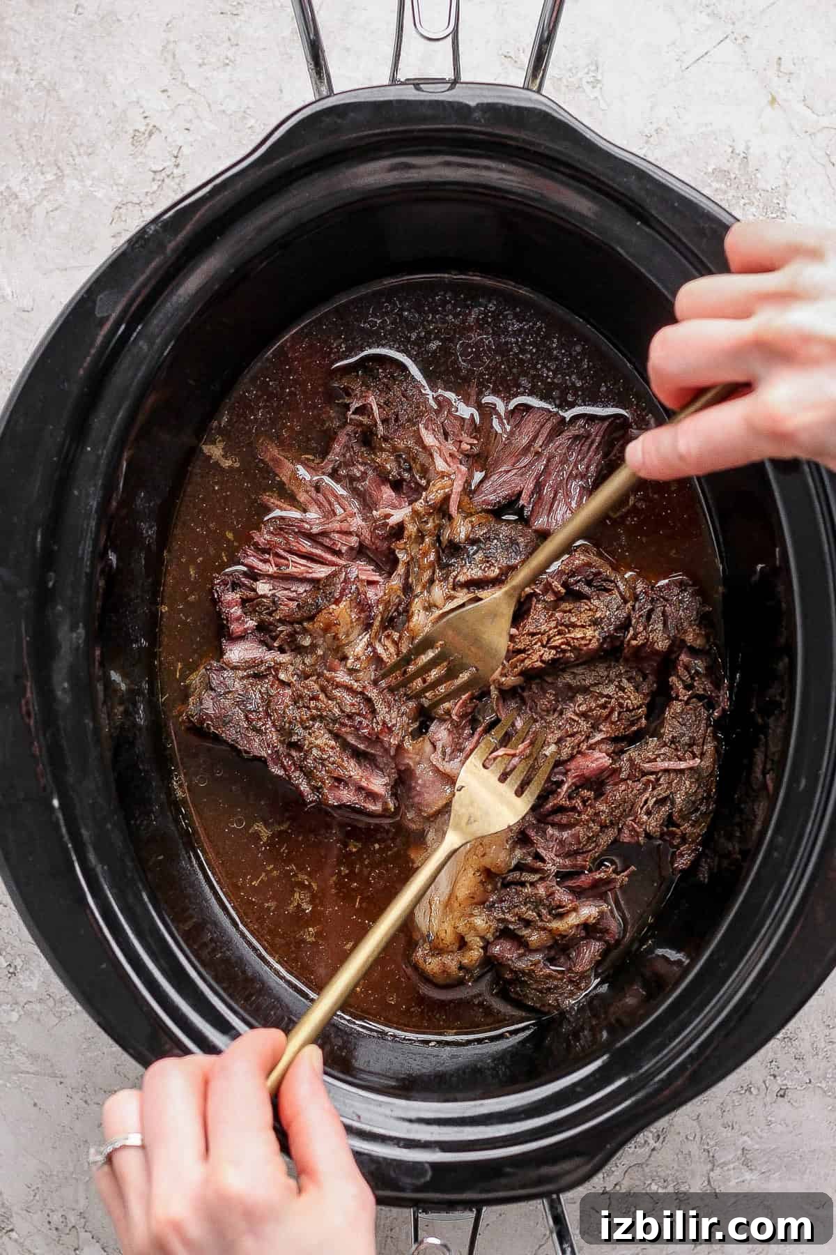 Two hands use forks to shred cooked beef inside a black slow cooker filled with broth on a light countertop.