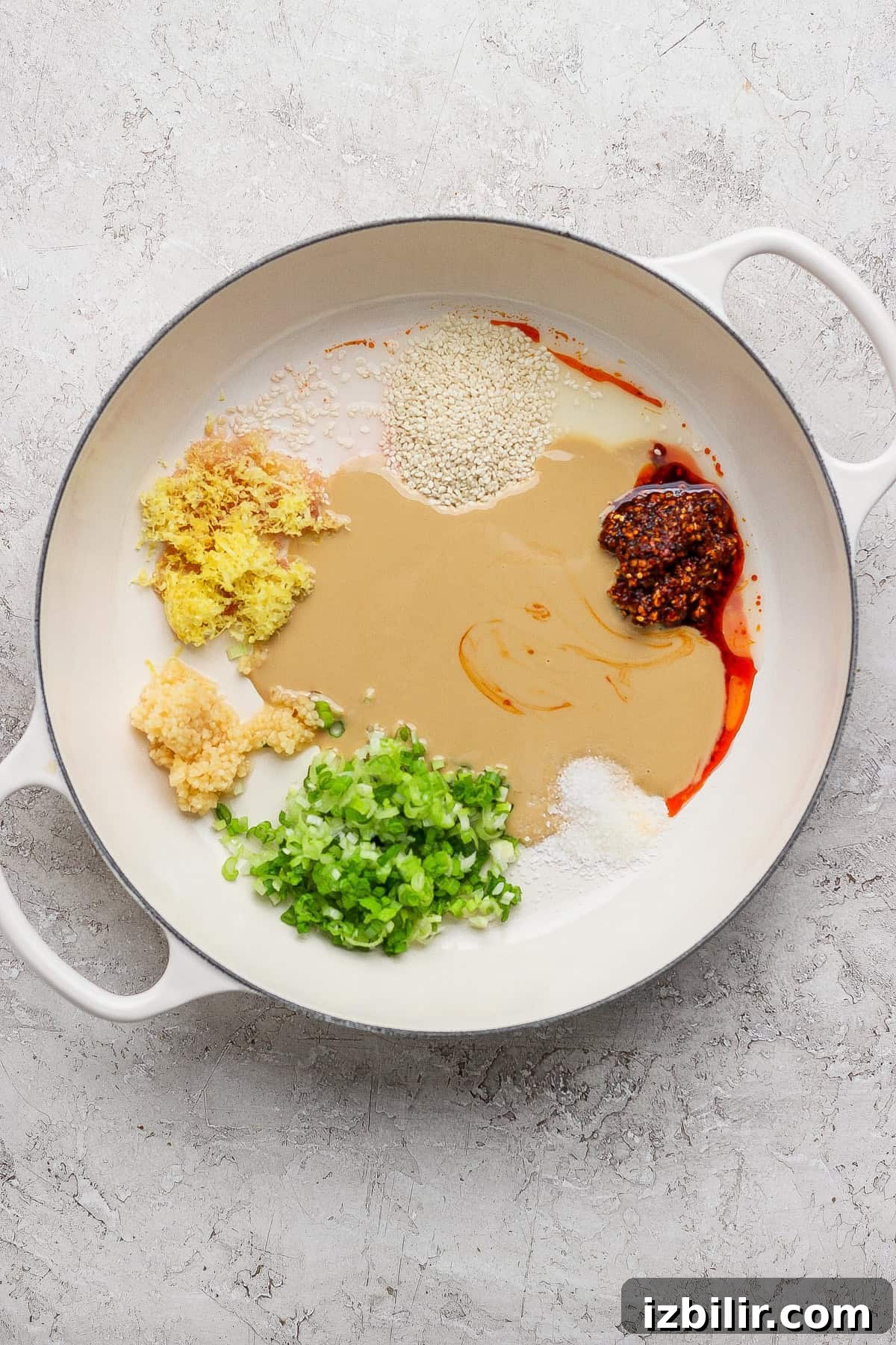 A white pan contains green onions, minced garlic, ginger, sesame seeds, tahini, chili oil, whole grain mustard, and sugar on a light textured surface.