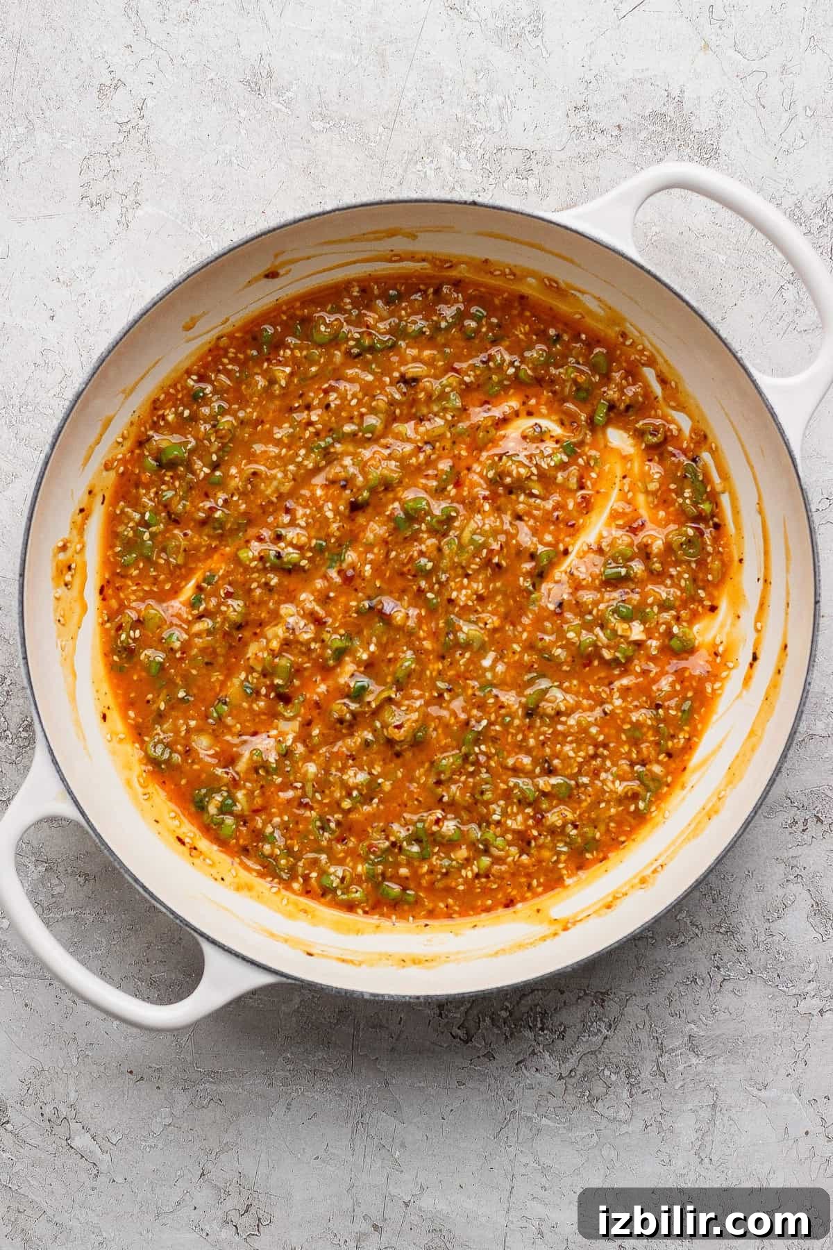A white pot filled with a thick, orange-red sauce mixed with chopped green herbs and visible seeds, sitting on a textured light surface.