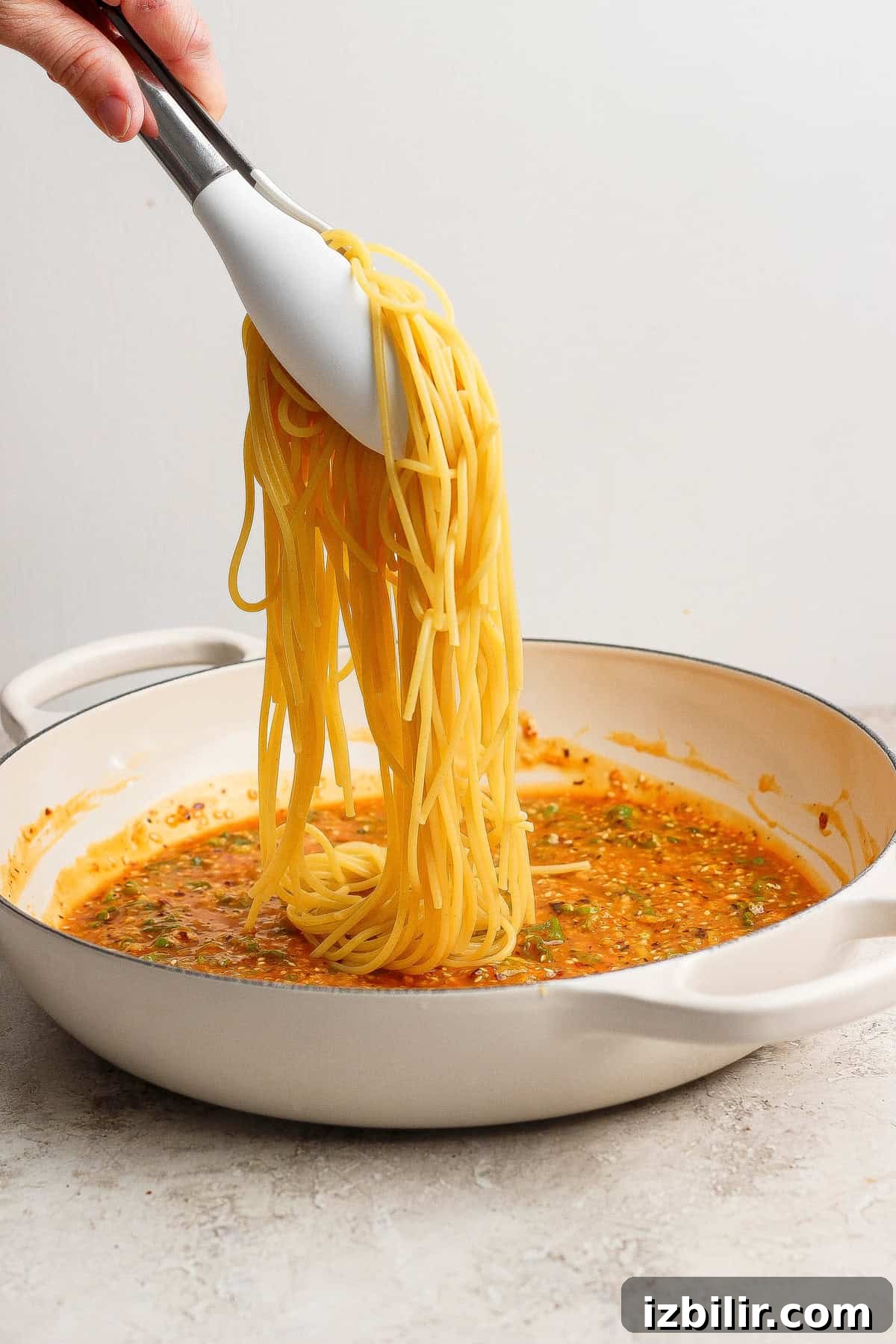 A hand uses tongs to lift cooked spaghetti over a white pan filled with tomato-based sauce on a light surface.
