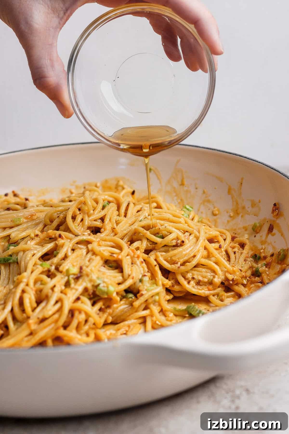 A hand pours liquid from a small glass bowl onto cooked spaghetti in a white pan.