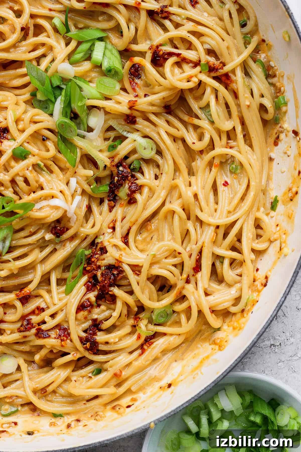 Cooked spaghetti coated in a creamy sauce, topped with chili flakes and chopped green onions, in a white pan with extra green onions in a small bowl nearby.