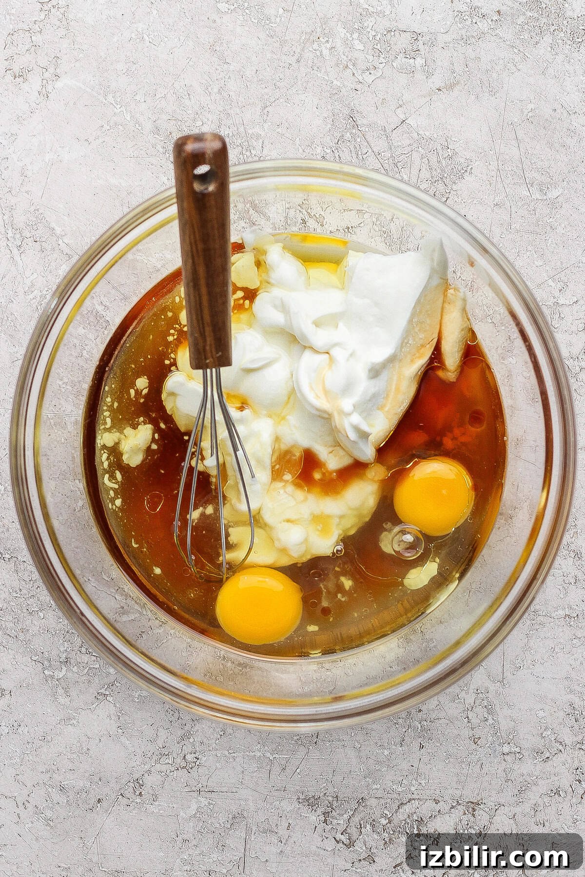 A glass bowl containing two raw eggs, yogurt, and a brown liquid with a metal whisk resting on the side, placed on a textured countertop.
