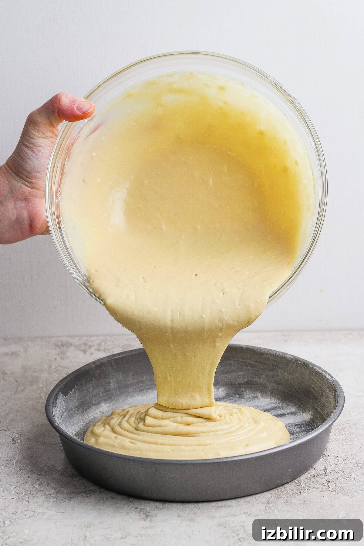 A hand pours cake batter from a glass bowl into a round, floured metal baking pan on a light countertop.