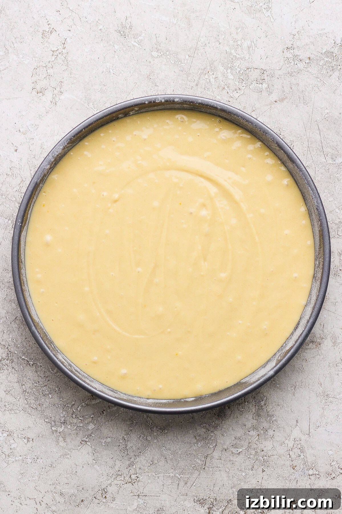 A round cake pan filled with smooth, pale yellow cake batter sits on a light textured surface.