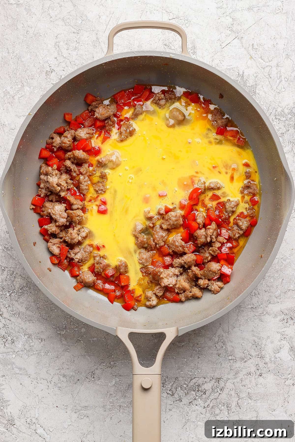 A skillet containing cooked sausage, diced red bell peppers, and beaten eggs beginning to cook.