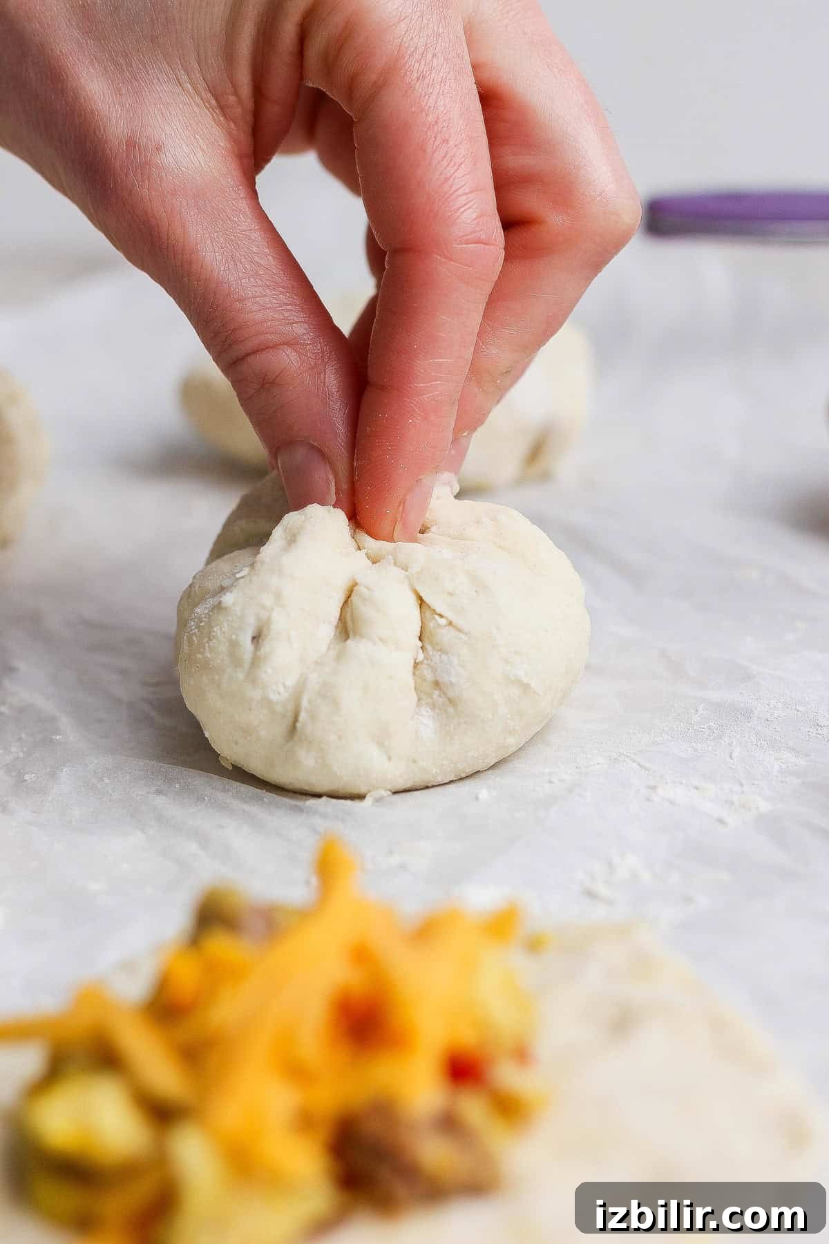 A hand pinches the top of a ball of dough to seal it closed; fillings and shredded cheese are visible on a piece of dough in the foreground.