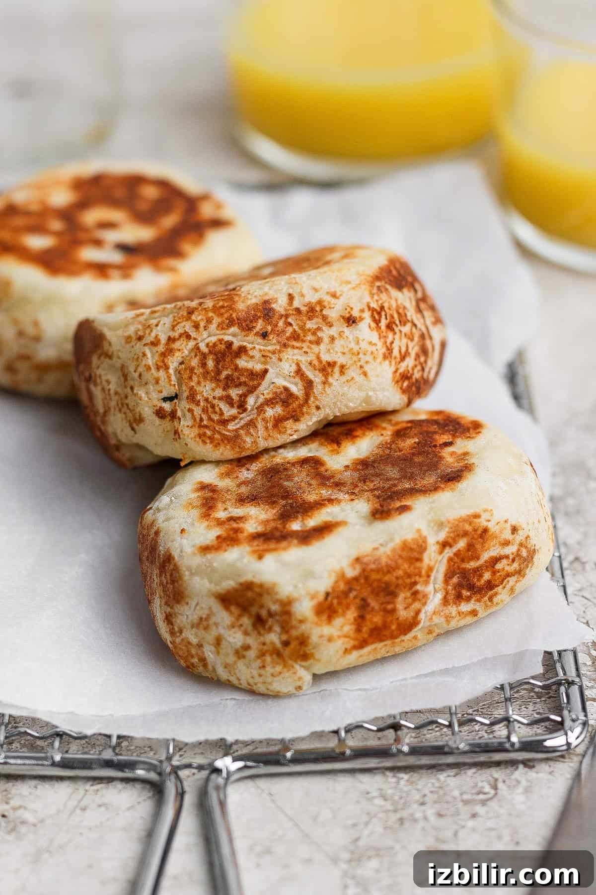 Three browned, thick English muffins rest on parchment paper atop a cooling rack, with blurred glasses of orange juice in the background.