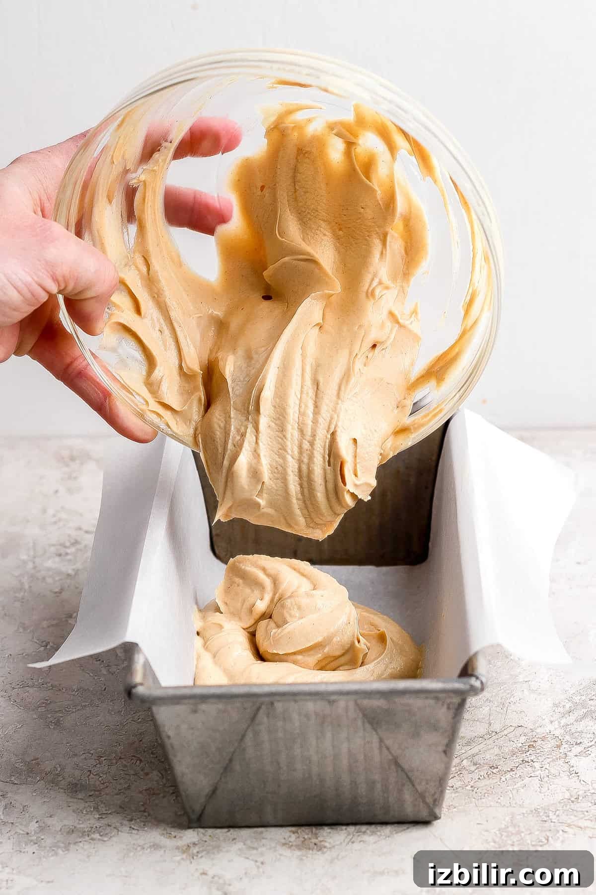Creamy Peanut Butter Frozen Yogurt Bites Recipe 4 A person pours thick, light brown batter from a glass bowl into a parchment-lined metal loaf pan on a light-colored surface.