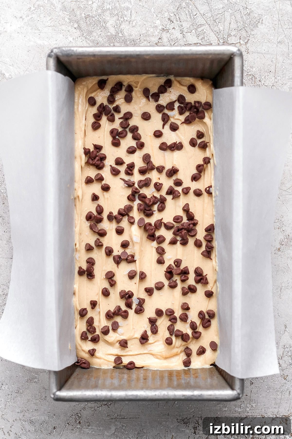 Creamy Peanut Butter Frozen Yogurt Bites Recipe 5 A rectangular metal loaf pan lined with parchment paper, filled with batter and topped with scattered chocolate chips, ready to be frozen.