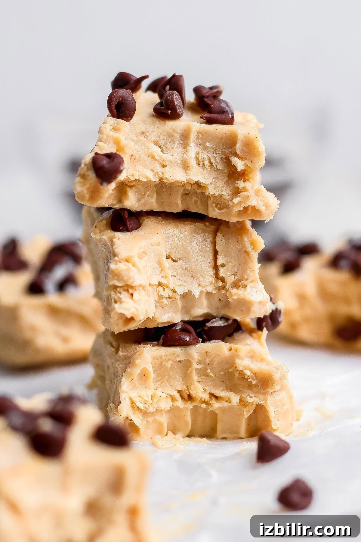 Creamy Peanut Butter Frozen Yogurt Bites Recipe 7 Three pieces of peanut butter fudge, stacked and topped with chocolate chips, with more pieces and chips scattered in the background.