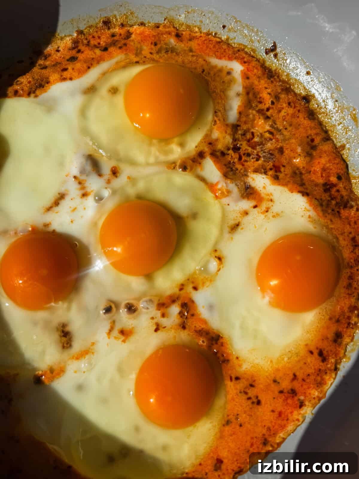 Five sunny-side-up eggs cooking in a pan, surrounded by orange-hued oil and spices.