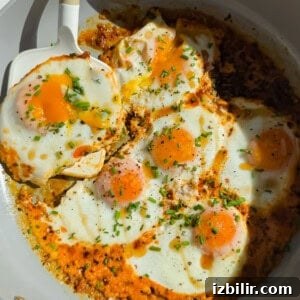 Six sunny-side up eggs cooking in a skillet with herbs and spices, one being lifted by a white spatula.