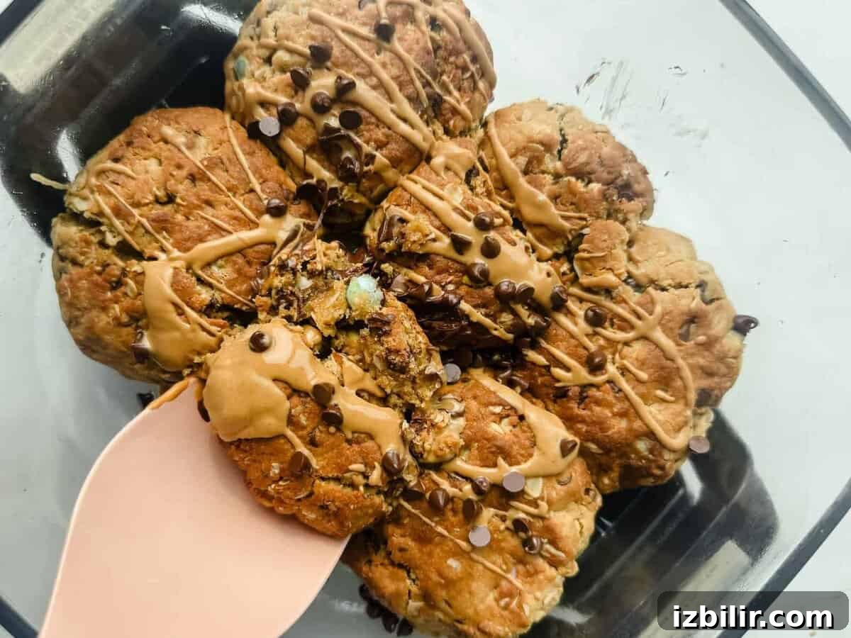 Scoopable Cookies: The Viral Recipe Everyone's Making 2 A glass dish containing seven chunky cookies drizzled with light brown icing and sprinkled with mini chocolate chips, one cookie being lifted with a pink spatula.