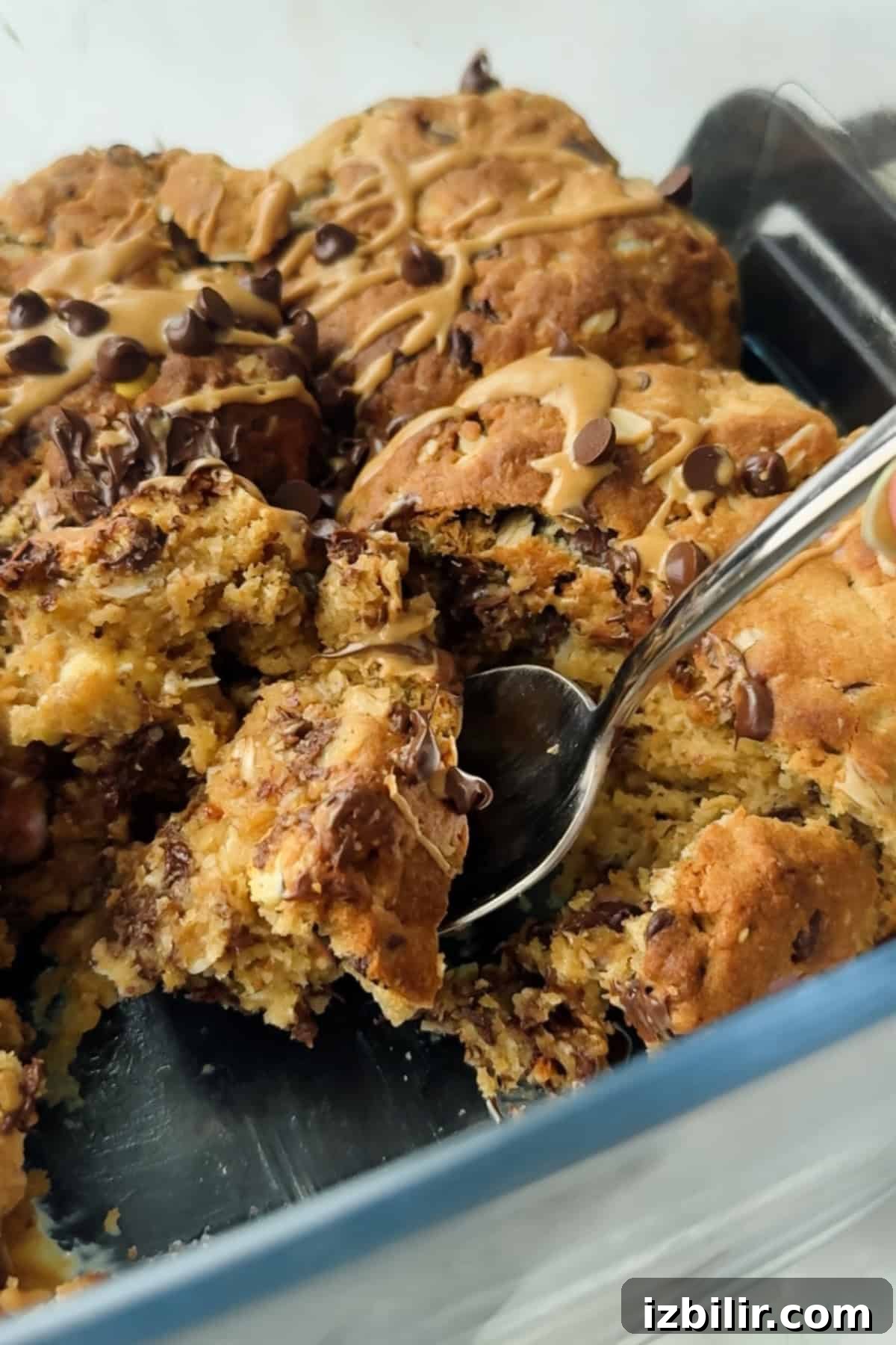 Scoopable Cookies: The Viral Recipe Everyone's Making 3 A close-up of a partially served chocolate chip cookie dessert in a glass dish, drizzled with a light glaze, with a spoon lifting a portion.