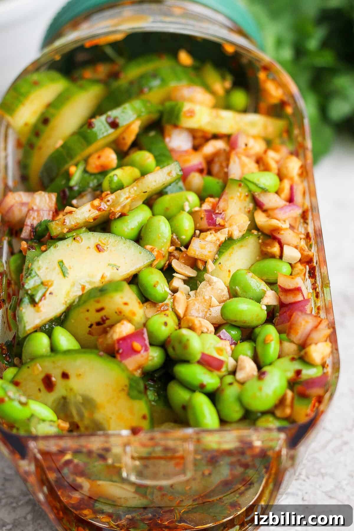 A close-up of a salad with sliced cucumbers, edamame, red onion, chopped nuts, and a red chili-flake dressing in a rectangular glass container.