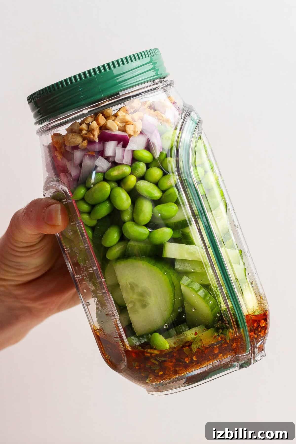 A hand holds a tilted plastic jar filled with layered ingredients including cucumber, edamame, red onion, walnuts, and dressing at the bottom.
