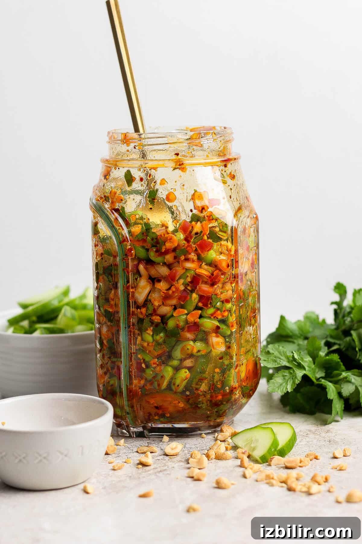 A glass jar filled with a colorful chopped vegetable salad, topped with sauce and a spoon, sits on a countertop with herbs, peanuts, and cucumber slices nearby.