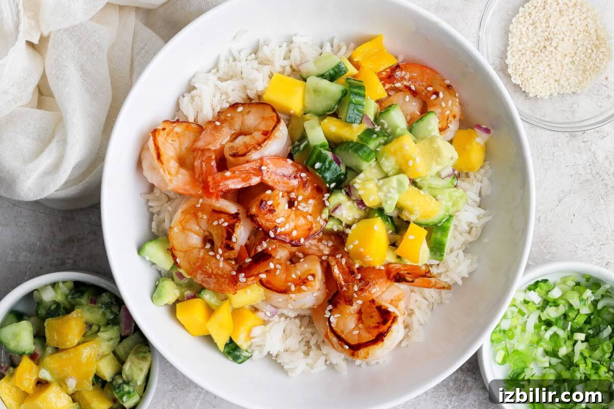 A bowl of white rice topped with grilled shrimp, diced mango, cucumber, avocado, and sprinkled sesame seeds, with side bowls of salsa and chopped green onions.