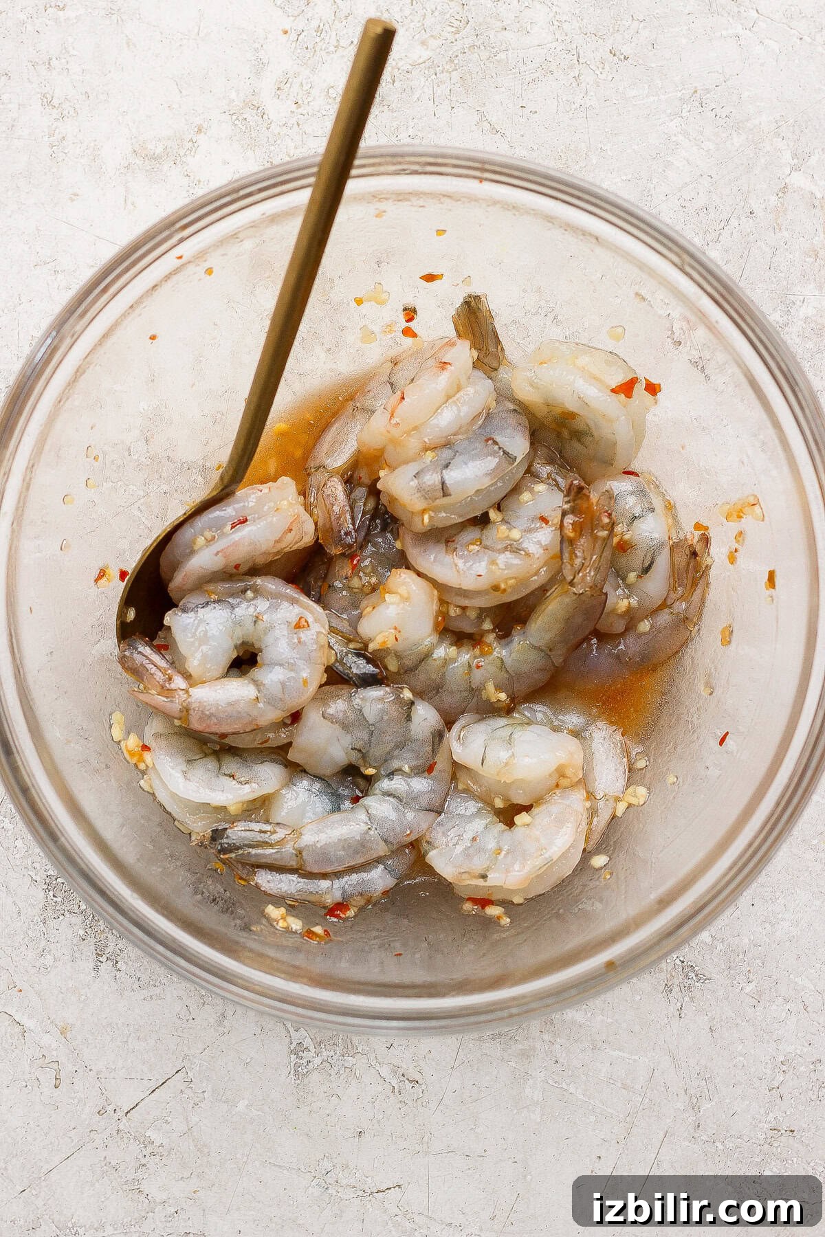 Raw shrimp marinating in a clear glass bowl with a metal spoon, mixed with a sauce containing red pepper flakes and minced garlic, on a light textured surface.