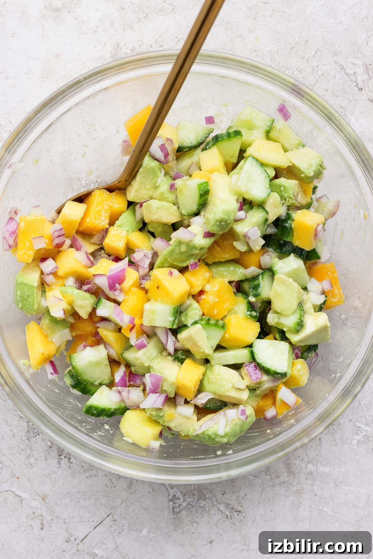 A glass bowl filled with chopped avocado, mango, cucumber, and red onion, mixed with a spoon on a light textured surface.