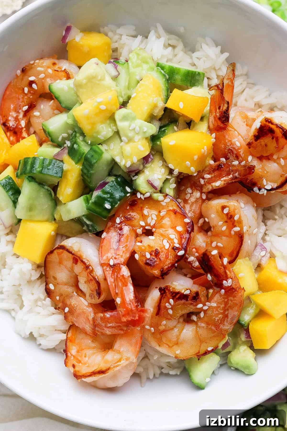A bowl of white rice topped with cooked shrimp, diced mango, avocado, cucumber, red onion, and sprinkled with sesame seeds.