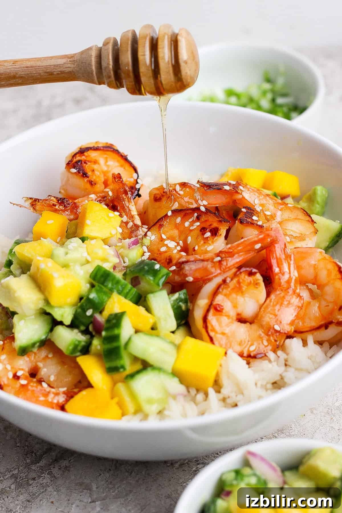 A bowl of rice topped with grilled shrimp, diced mango, cucumber, avocado, and sesame seeds, with honey being drizzled on top.