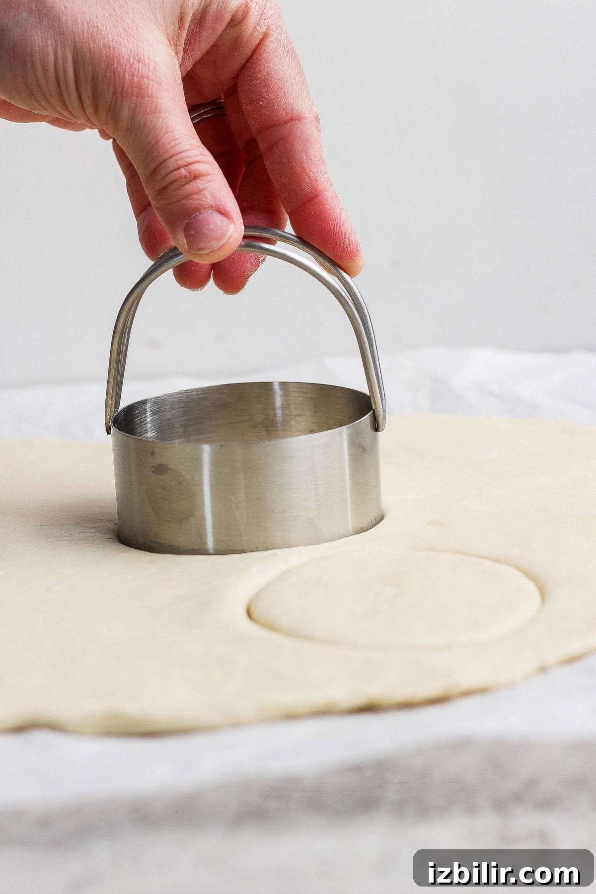 A hand presses a round metal biscuit cutter into rolled-out dough on a flat surface.