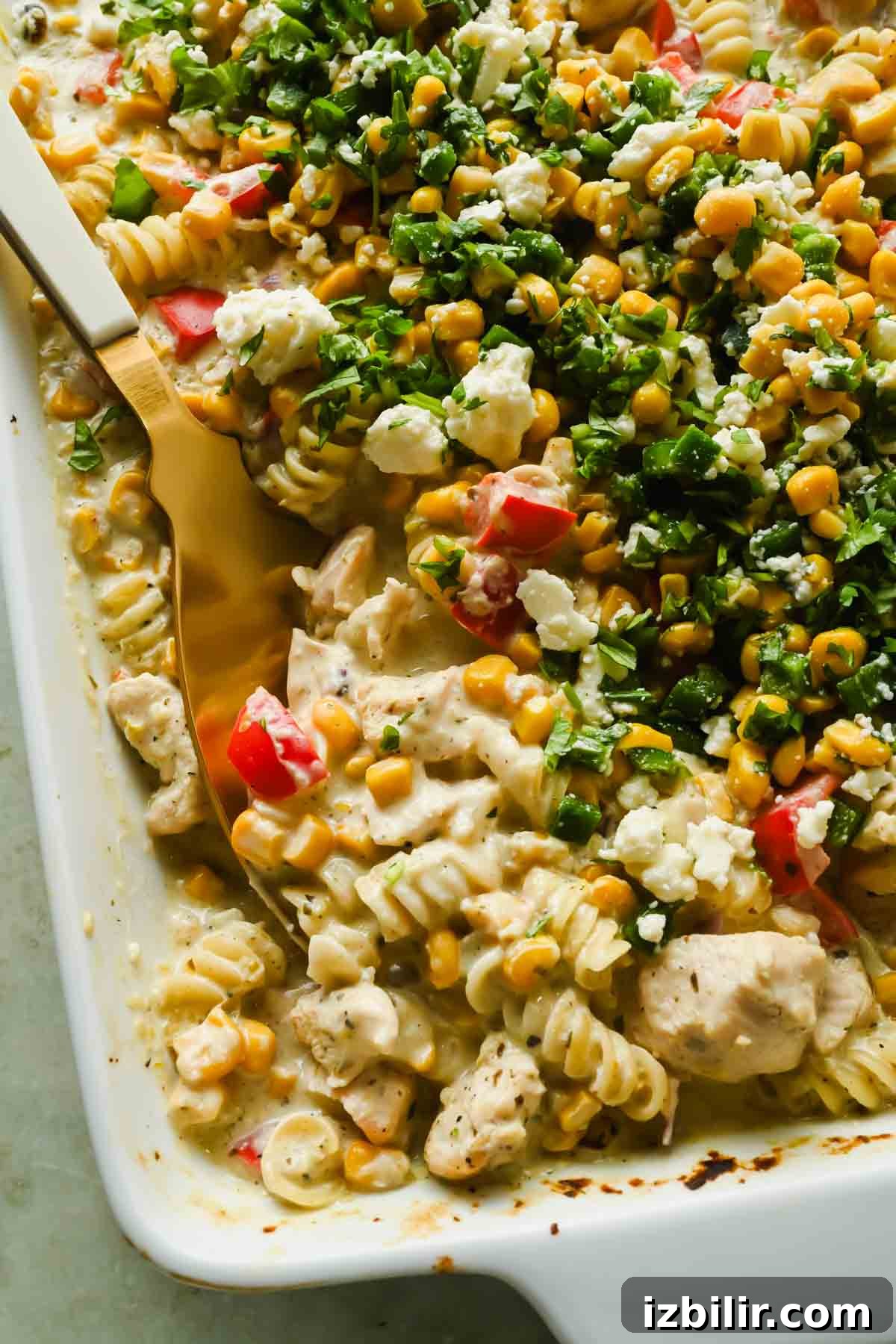 Creamy Oven Baked Street Corn Pasta with Cotija 5 Baked pasta casserole with rotini, diced chicken, corn, red bell peppers, creamy sauce, and fresh chopped herbs, served in a white baking dish with a gold serving spoon.