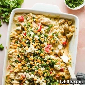Creamy Oven Baked Street Corn Pasta with Cotija 7 Baking dish with creamy chicken, corn, chopped red peppers, green herbs, and cheese, topped with chopped cilantro and cheese; a serving spoon is inside the dish.