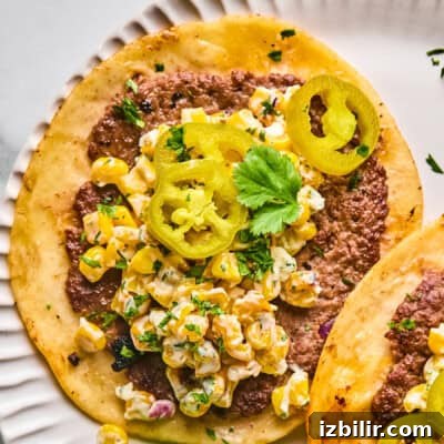 A close-up of a taco with ground beef, creamy corn salad, pickled jalapeño slices, and a sprig of cilantro on a white plate.