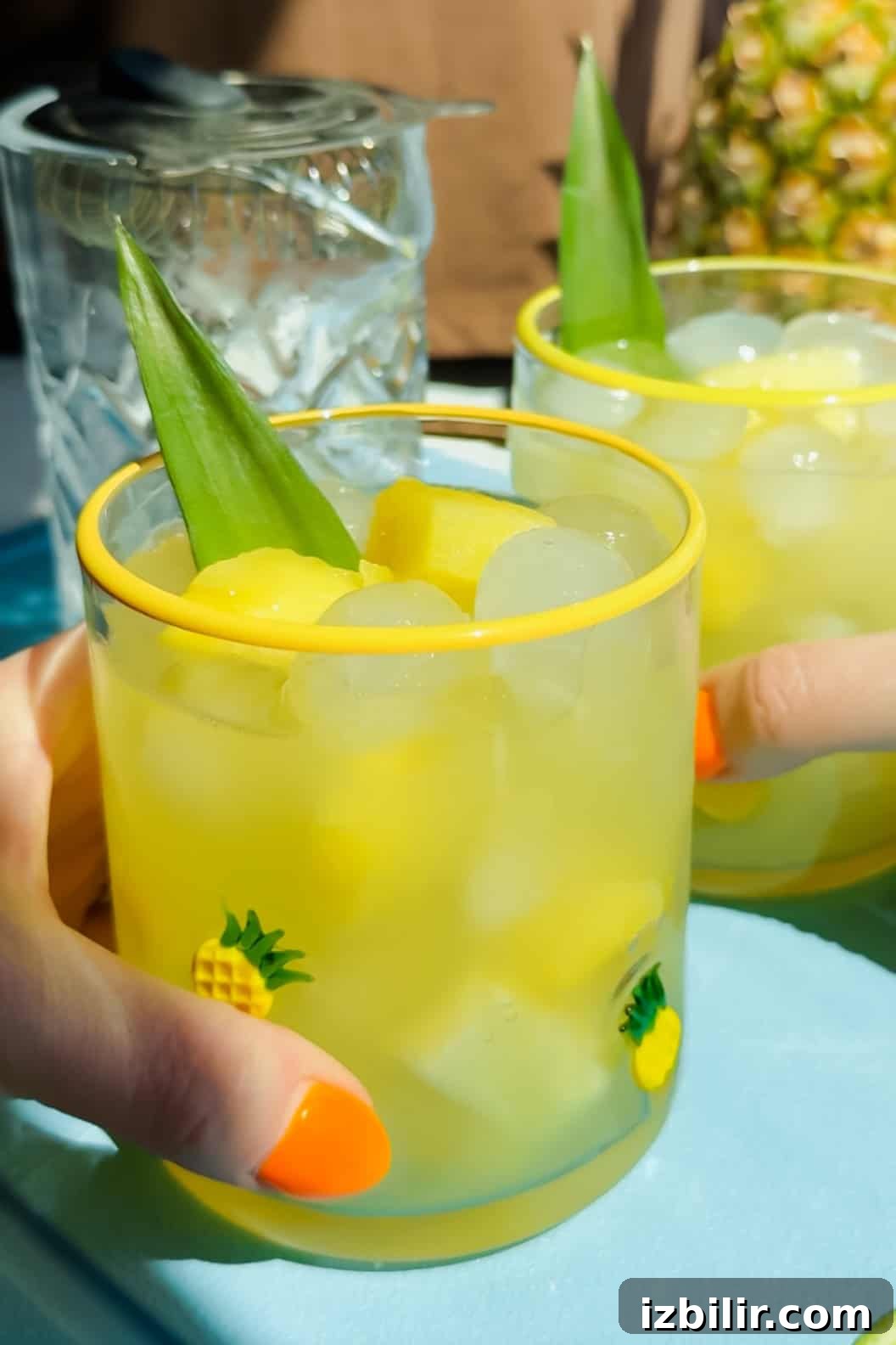 Two hands holding glasses filled with ice, yellow drink, pineapple chunks, and a pineapple leaf garnish, decorated with small pineapple designs.