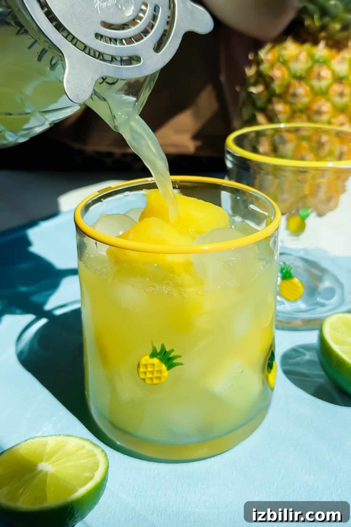 A glass with ice and yellow frozen fruit is being filled with juice. The glass has pineapple designs. Lime halves and another glass are nearby.